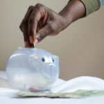 Close-up of a person's hand placing coins into a transparent piggy bank to save money.